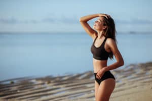 Outdoor shot of smiling young female model in bikini standing against blue sky. Woman having fun out on a summer day.