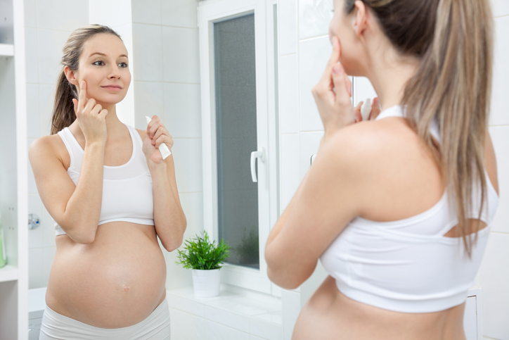 Pregnant woman applying lotion to face while looking in the mirror